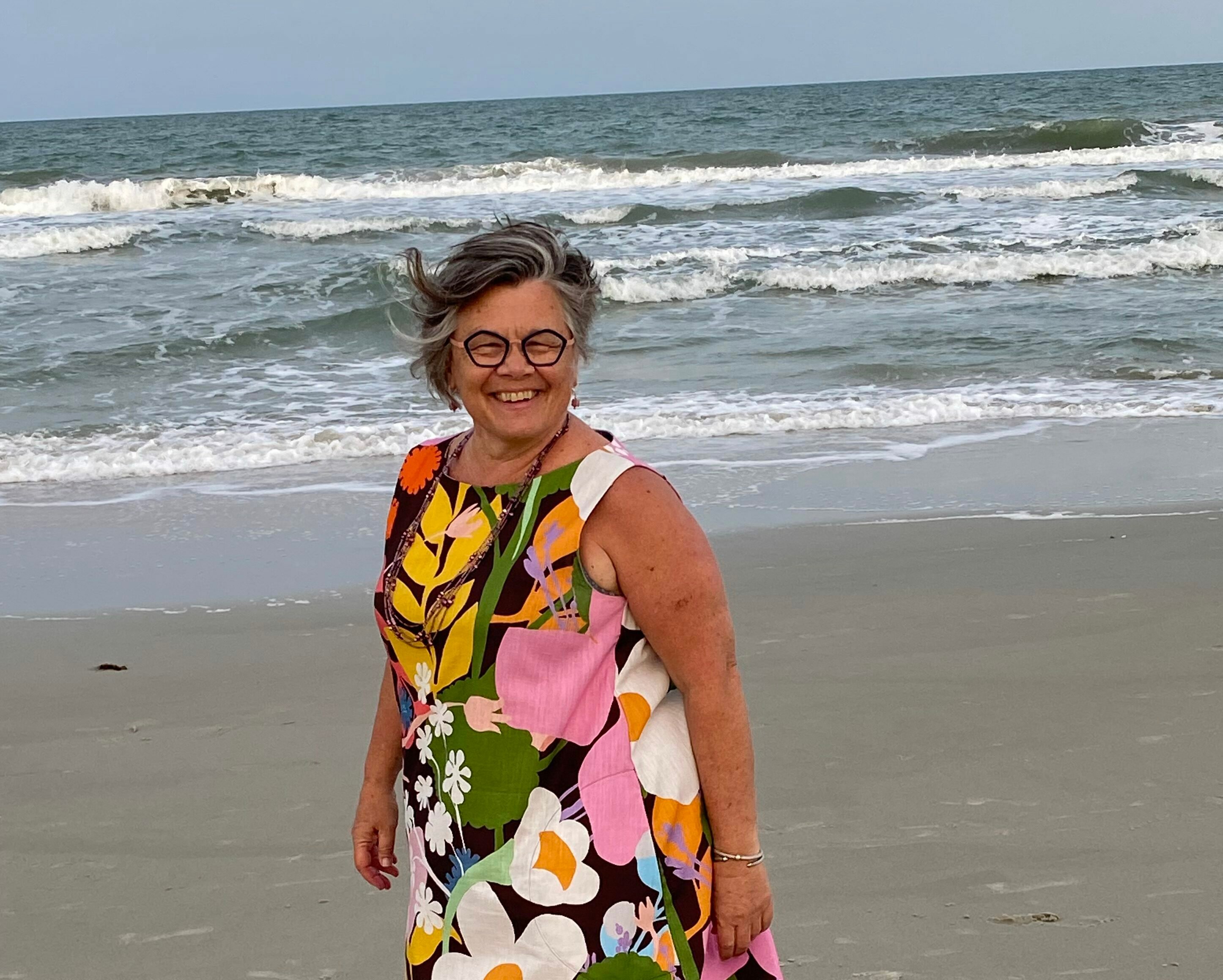 Tamela Rich on the beach with the wind in her hair. She's wearing a dress with big flowers and her eyeglasses.