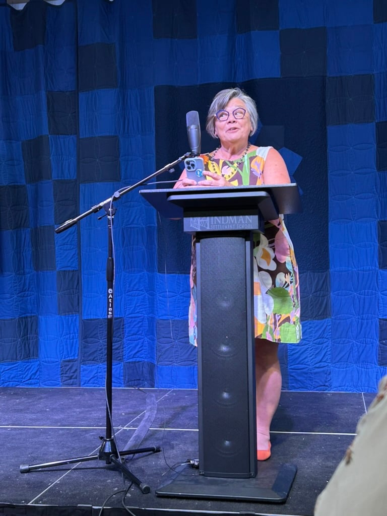 Woman standing behind a lecture reading from her phone at the microphone, wearing glasses and a colorful dress. A blue quilted backdrop